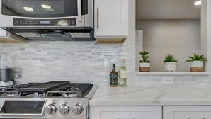 Een moderne keuken met roestvrijstalen apparatuur, een magnetron en een gasfornuis. Op het aanrecht staan drie flessen en erboven een plantenplank.