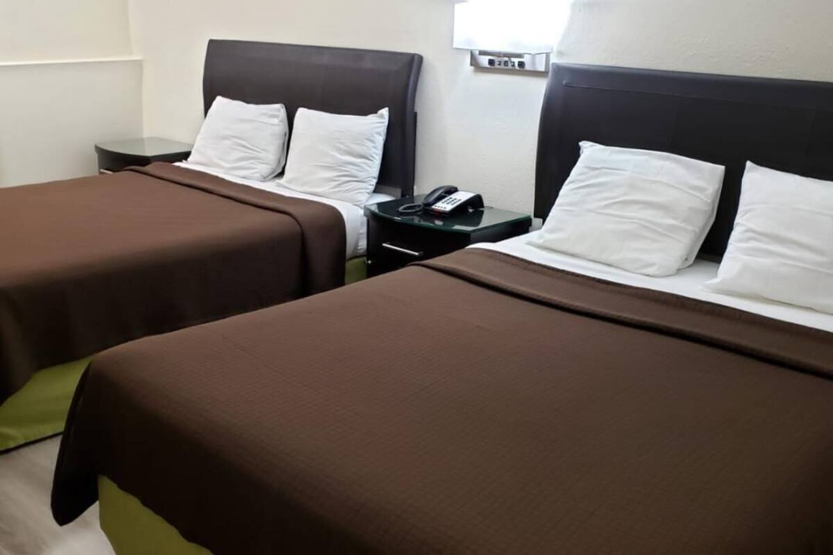 A hotel room with two beds, each with a brown bedspread and white pillows, a bedside table with a phone, and a wall-mounted light.