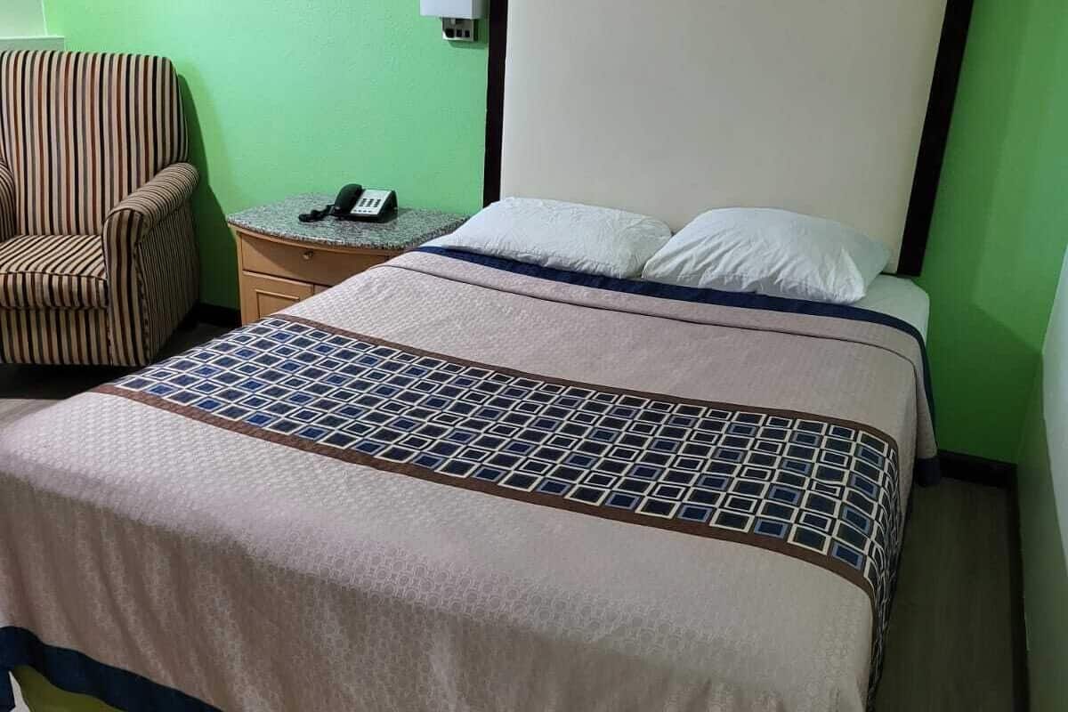 A hotel room with a bed, a striped armchair, a bedside table with a phone, and a wall-mounted air conditioner.