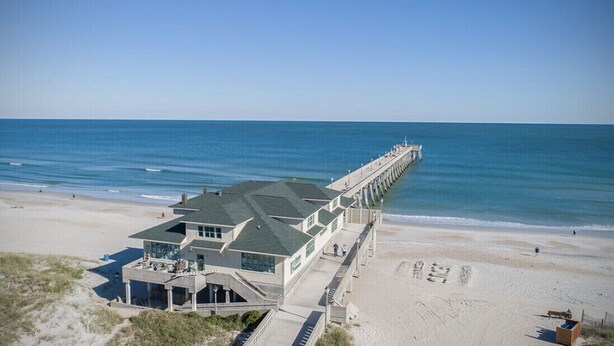 A beachfront property with a long pier extending into the ocean, a large building with multiple windows, and a sandy beach area.