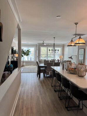 A modern kitchen with a dining area, featuring a large island, pendant lighting, and a view of the outdoors through large windows.