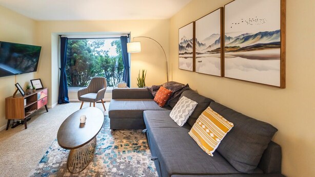 Ein modernes Wohnzimmer mit grauem Sofa, einem Couchtisch und einem an der Wand montierten Fernseher. Über dem Sofa befinden sich gerahmte Kunstwerke und es gibt ein Fenster mit Vorhängen.