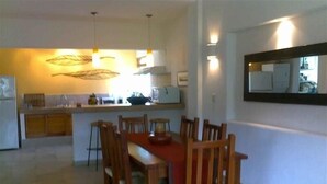 A dining area with a wooden table and chairs, a kitchen in the background, and a mirror on the wall.