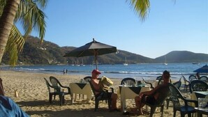 A beach with tables and chairs under umbrellas, a clear sky, and boats in the water.