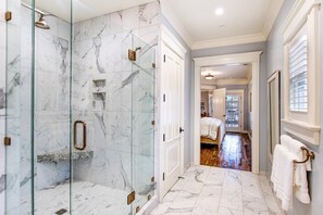 A modern bathroom with a glass-enclosed shower, marble flooring, and a hallway leading to a bedroom.