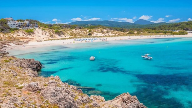 Una playa costera con aguas turquesas cristalinas, terreno rocoso y un edificio con varios balcones con vista a la playa.