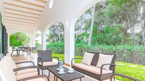 Un patio al aire libre con mobiliario de mimbre, una mesa con vasos y una jarra, y vistas a un exuberante jardín.