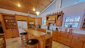 A kitchen with wooden cabinets, a central island with stools, a microwave, a stove, and a window with a view of trees. - Fox Den- MAGNIFICENT Lake View! 5 minute walk or drive to shoreline!  (Garfield)