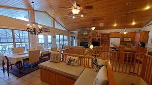 A spacious living area with a high ceiling, wooden flooring, a central seating area with a sofa and chairs, a dining table, and a kitchen area with wooden cabinets and a stone fireplace. - Fox Den- MAGNIFICENT Lake View! 5 minute walk or drive to shoreline!  (Garfield)