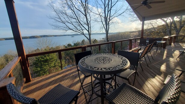 A covered outdoor deck with a round table and chairs, overlooking a lake with trees and a distant shoreline. - Fox Den- MAGNIFICENT Lake View! 5 minute walk or drive to shoreline!  (Garfield)