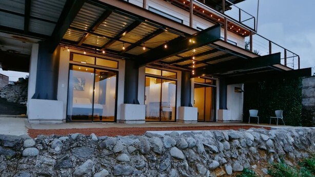 A modern house with a stone retaining wall, large glass doors, and a covered porch area. - Cozy Retreat with Breathtaking Views, 20 Minutes to the Airport (Alajuela)