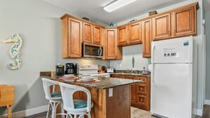 Una cocina con armarios de madera, un microondas, un refrigerador y una zona de bar con dos sillas blancas.