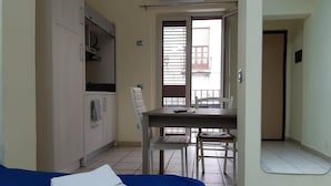 Ein kleiner, gut beleuchteter Raum mit einem Bett, einem Tisch mit Stühlen, einer Küchenzeile und einem Fenster mit Jalousien.
