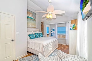 Un dormitorio con cama, ventilador de techo, televisión y vista a la playa a través de una ventana.