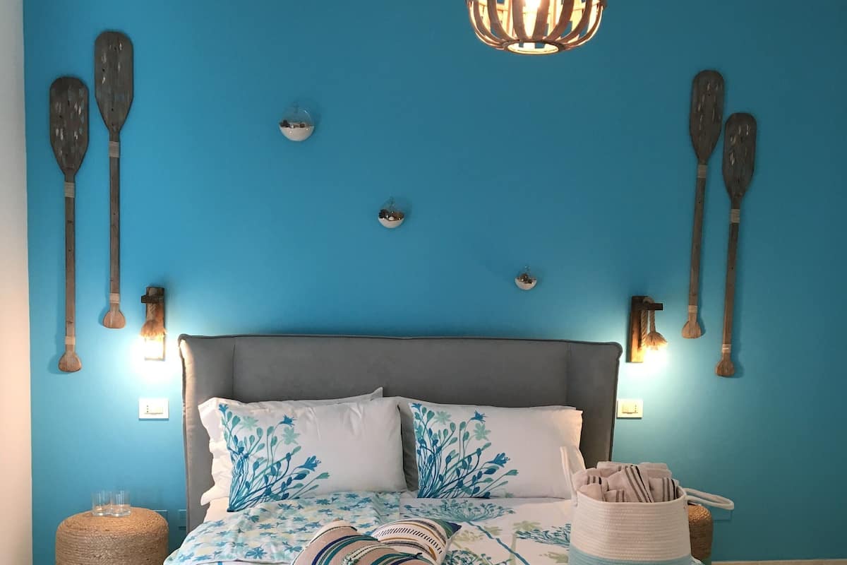 A bedroom with a blue wall, a grey headboard, and a bed with a striped bedspread. There are two wall-mounted lights and several wooden paddles on the wall.