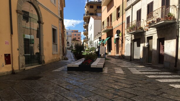 Una calle estrecha con pavimento de piedra, edificios con balcones y una jardinera central con flores.