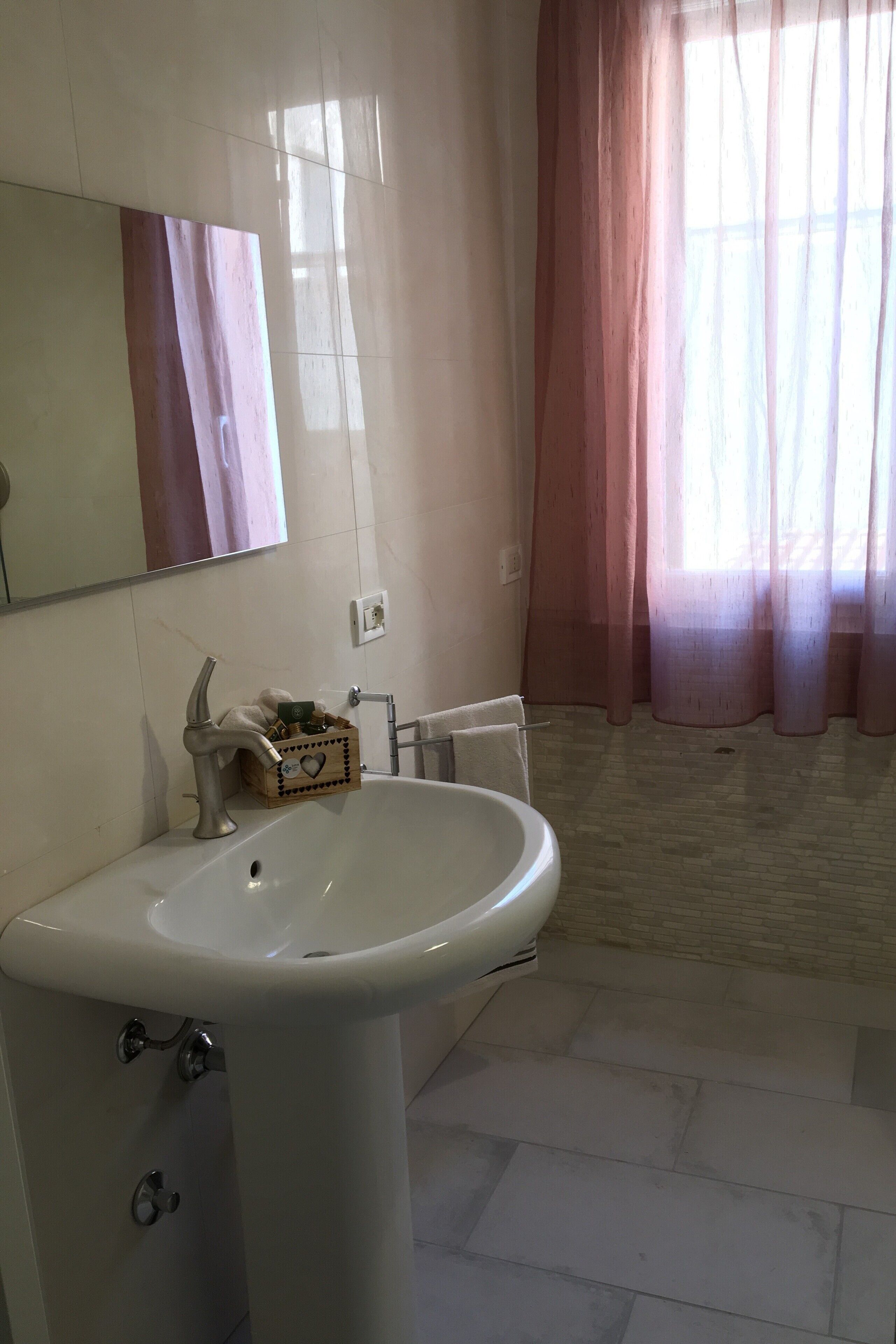 Un baño con lavamanos blanco, espejo, toallero y ventana con cortinas de gasa.