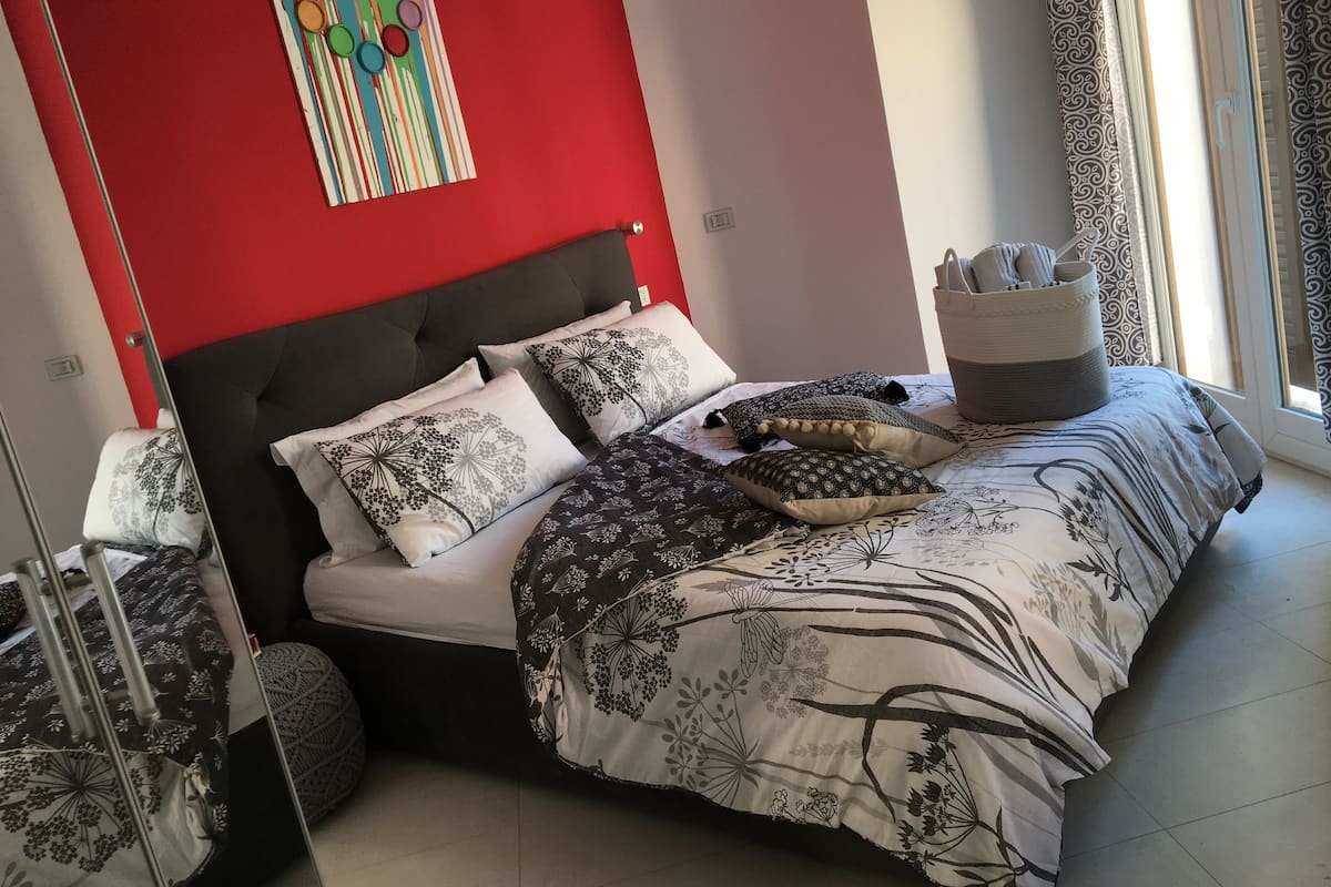 A bedroom with a bed, pillows, a bedside table, a wall art piece, and a window with curtains.