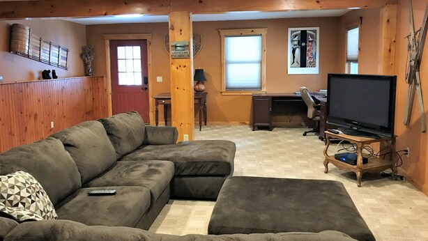 A living room with a sectional sofa, a TV on a stand, and a wooden accent wall. - Beautiful chalet with spectacular views in the Adirondack Mountains (Old Forge)