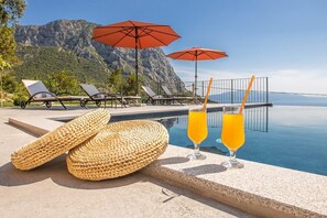 A poolside area with lounge chairs, two umbrellas, and two glasses of orange juice.