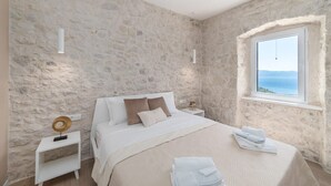Un dormitori amb una paret de pedra, un llit amb roba de llit blanca, una taula de nit amb una làmpada i una finestra amb vista al mar.
