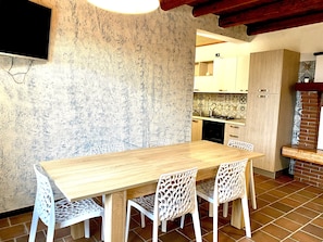 A dining area with a wooden table and white chairs, a kitchen in the background, and a TV mounted on the wall.