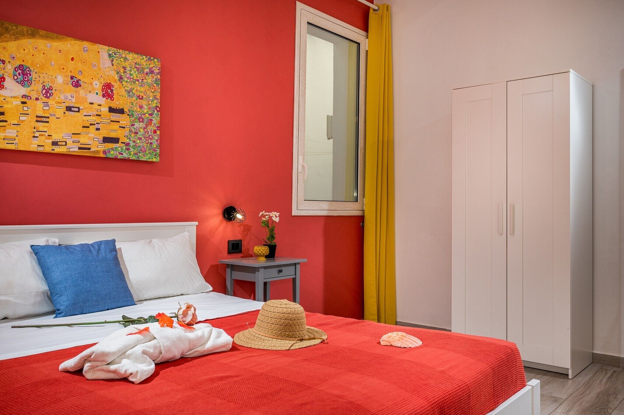 A hotel room with a red bedspread, a white headboard, a painting on the wall, a small table with a plant, and a wardrobe.