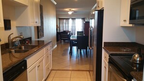 A kitchen with a dishwasher, sink, and refrigerator. There is a dining area with a table and chairs in the background. - Dawn 1024 (Galveston)