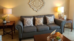 A living room with a grey sofa, patterned cushions, a decorative wall piece, and two side tables with lamps and plants. - Dawn 1024 (Galveston)