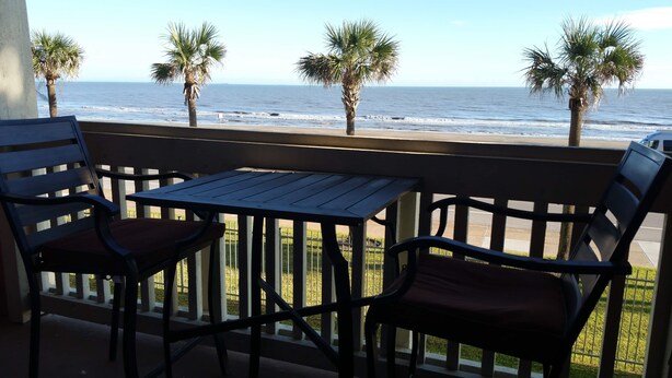 A balcony with a table and two chairs, overlooking the ocean with palm trees in the background. - Dawn 1024 (Galveston)