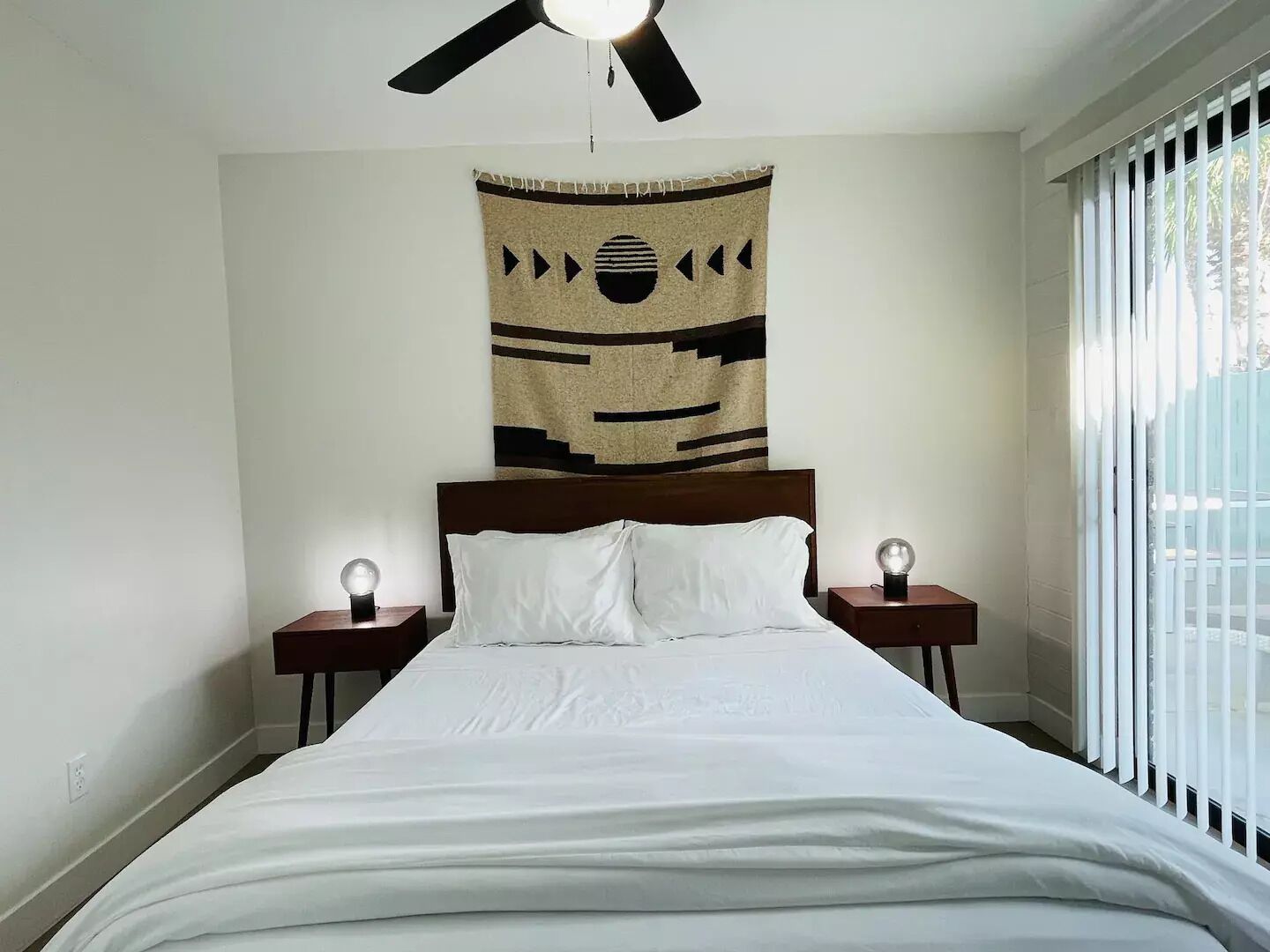 A bedroom with a bed, two bedside lamps, a ceiling fan, and a window with blinds.
