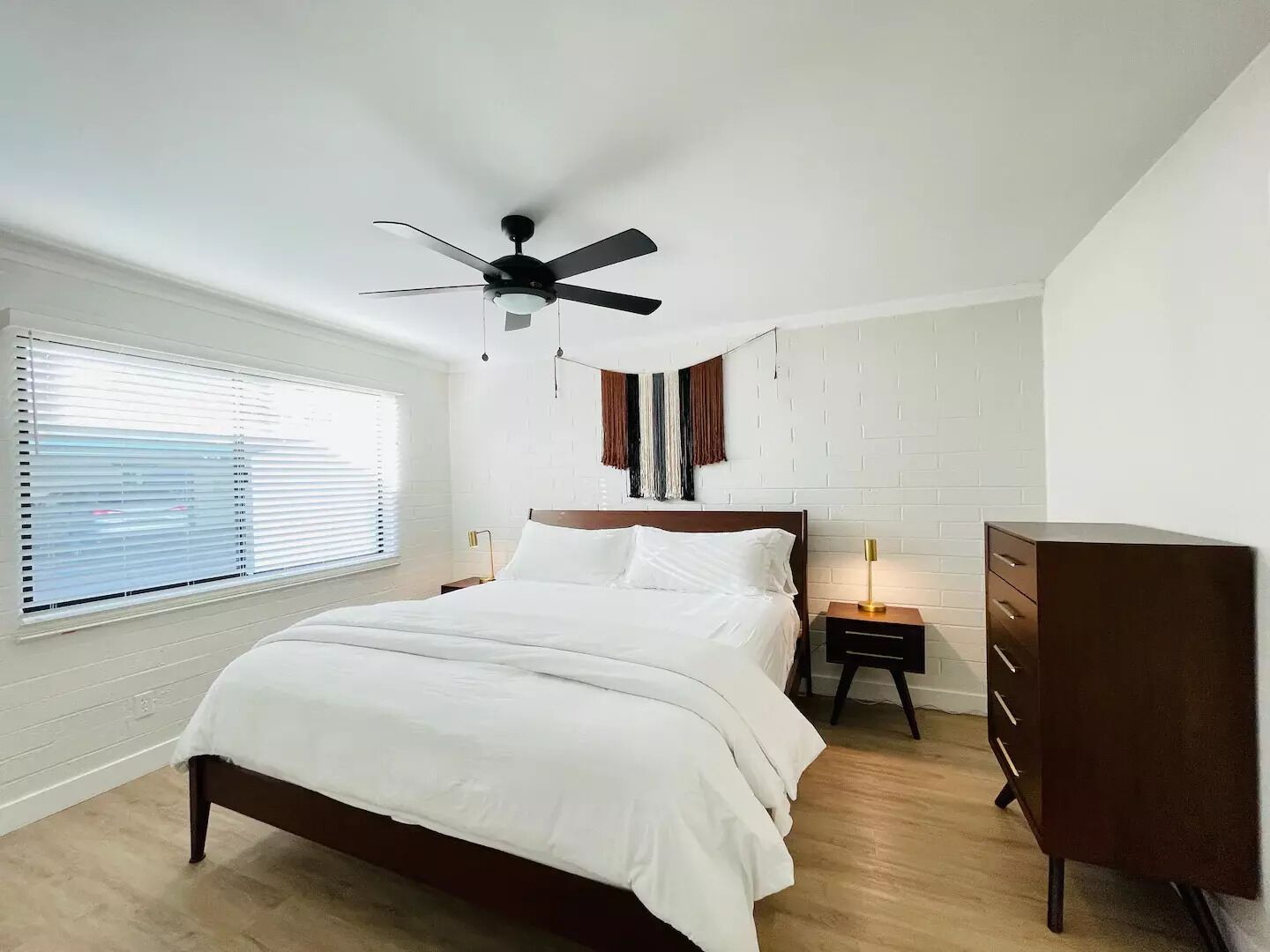Ein Schlafzimmer mit Bett, Kommode, Deckenventilator und einem Fenster mit Jalousien.