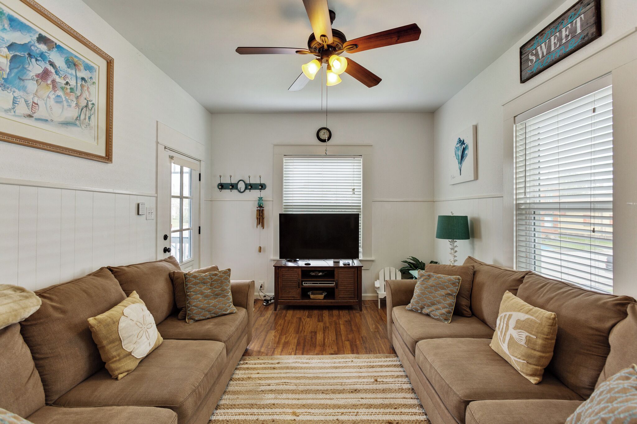 A living room with two brown sofas, a ceiling fan, a flat-screen TV, and a large painting on the wall.