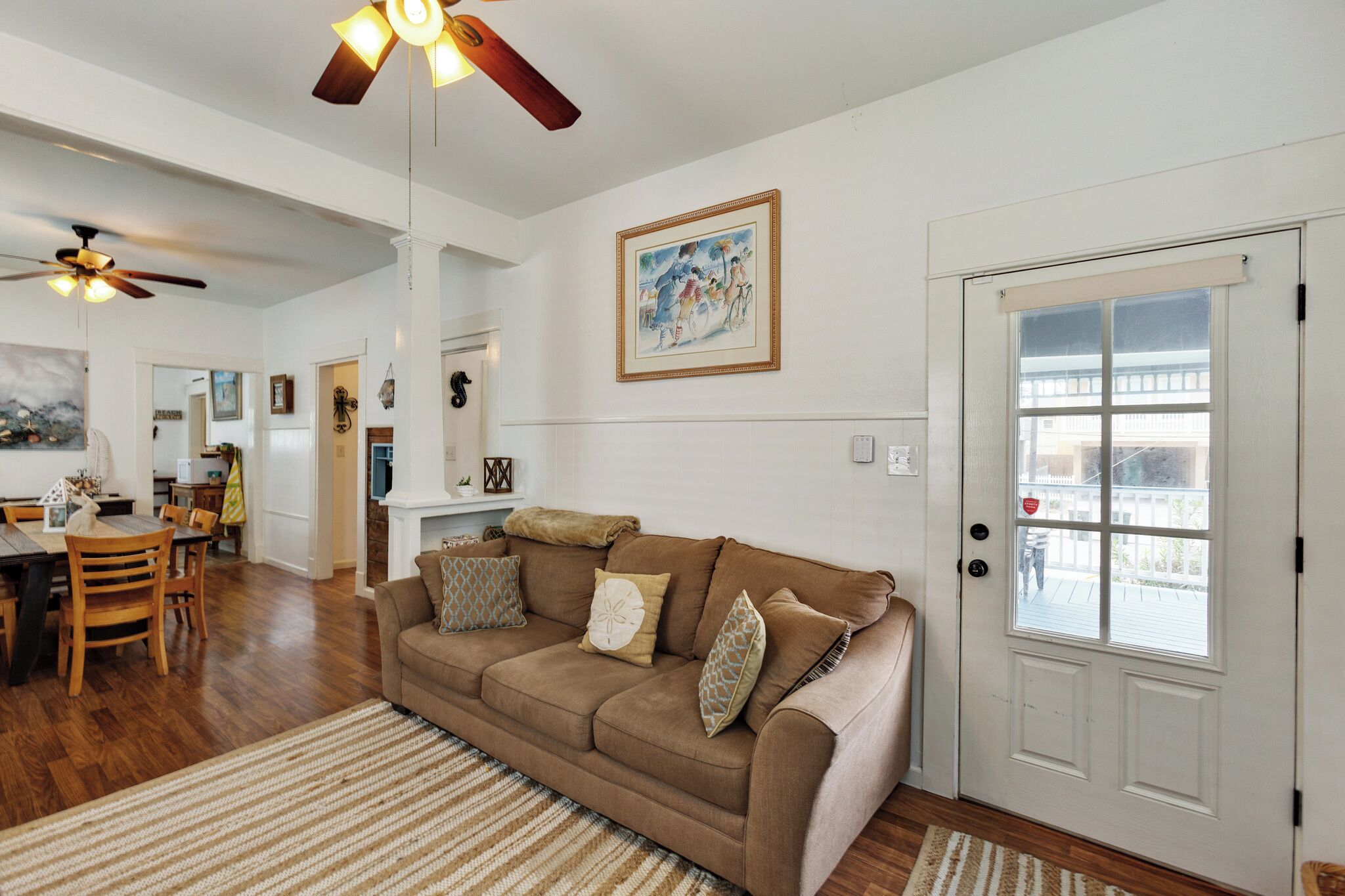 A living room with a brown sofa, wooden dining table, and chairs. There is a ceiling fan, a door with a window, and a framed picture on the wall.