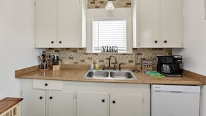 A kitchen with white cabinets, a double sink, a coffee maker, and a window with blinds. - Tina Marie (Galveston)