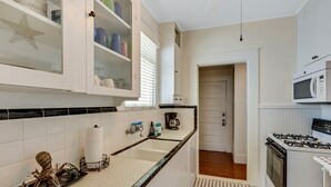 Cocina con armarios blancos, microondas y estufa. Se observa una ventana con persianas, un lavamanos y una cafetera en la encimera.