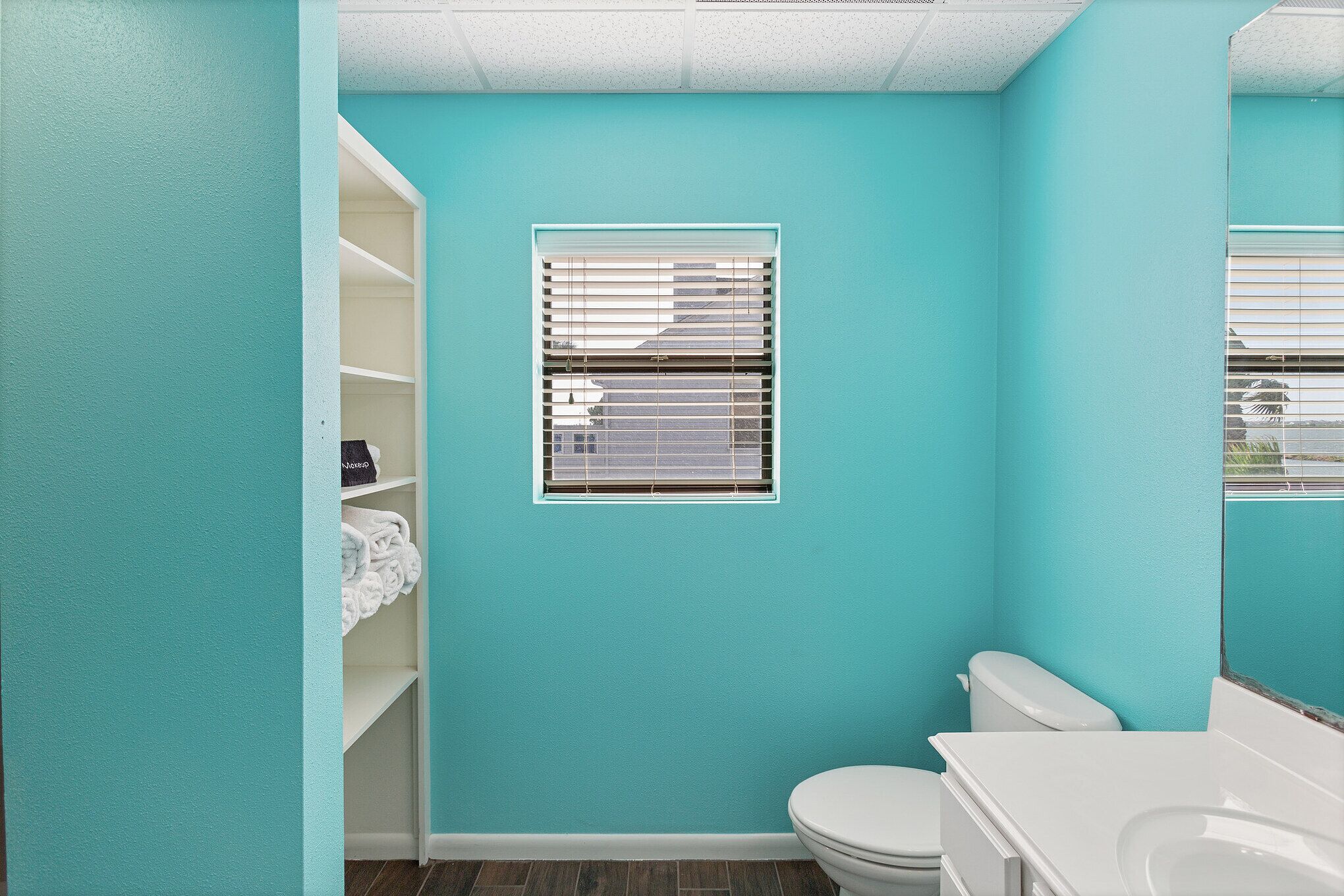 A bathroom with teal walls, a white toilet, a white sink, and a window with blinds.