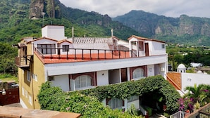 Il s’agit d’un bâtiment à deux étages, doté d’un toit recouvert de tuiles rouges, de murs blancs et d’un balcon. Il est entouré d’une végétation luxuriante et on aperçoit une montagne imposante en arrière-plan.
