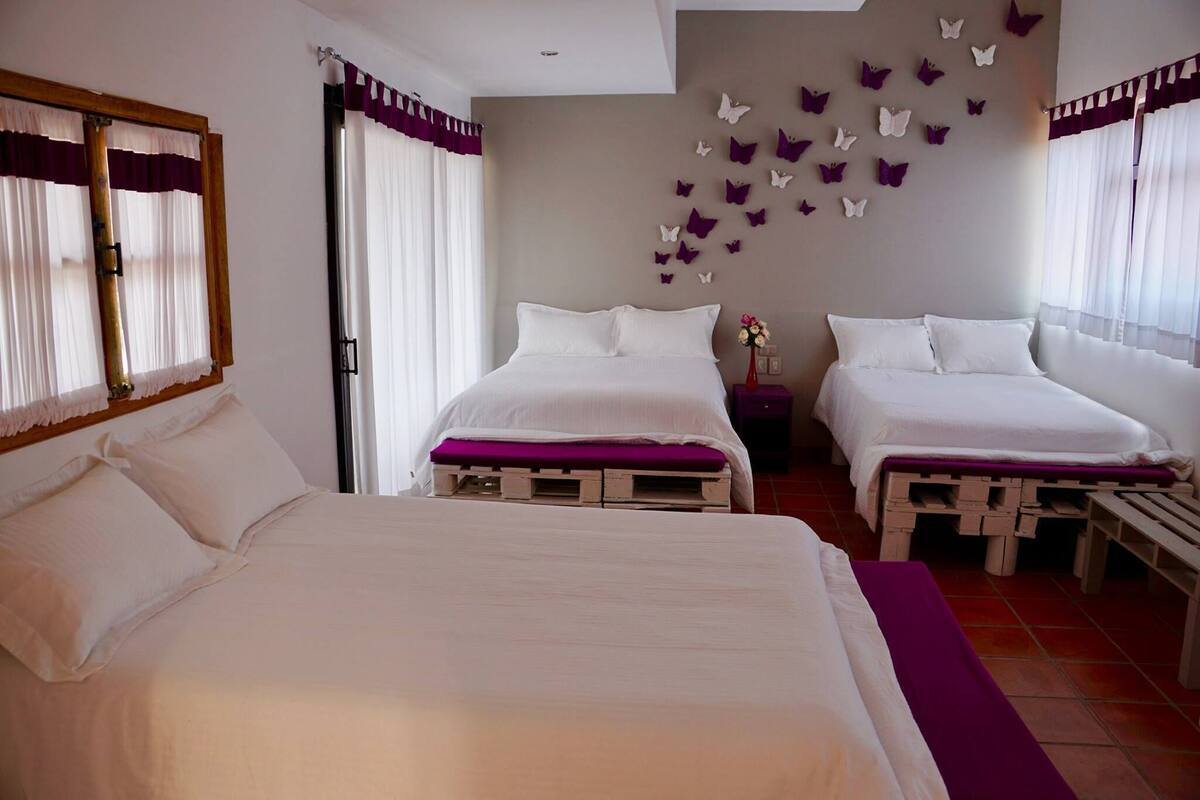 Hotellihuone, jossa on kaksi sänkyä, puinen pääty, violetti sängynpäällinen, valkoiset vuodevaatteet, perhoskoristein koristeltu seinä ja ikkuna, jossa on verhot.