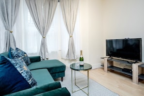 A modern living room with a green sofa, a small round table with a bottle and glass, and a flat-screen TV on a wooden stand.