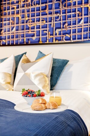 A neatly made bed with a white and blue pillow arrangement, a plate with croissants, a bowl of fresh berries, and a glass of orange juice.