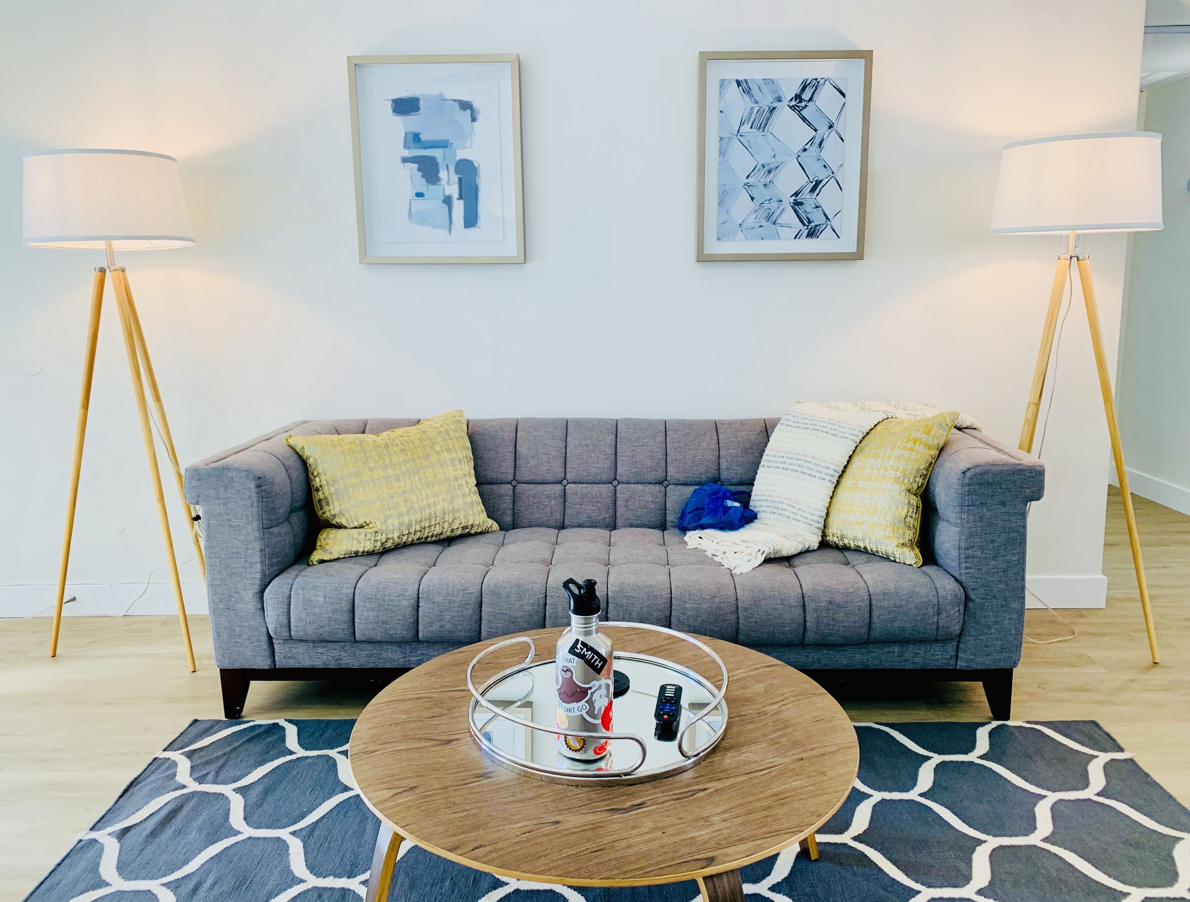 A living room with a grey sofa, two framed abstract artworks, a round coffee table with a tray, and two floor lamps.