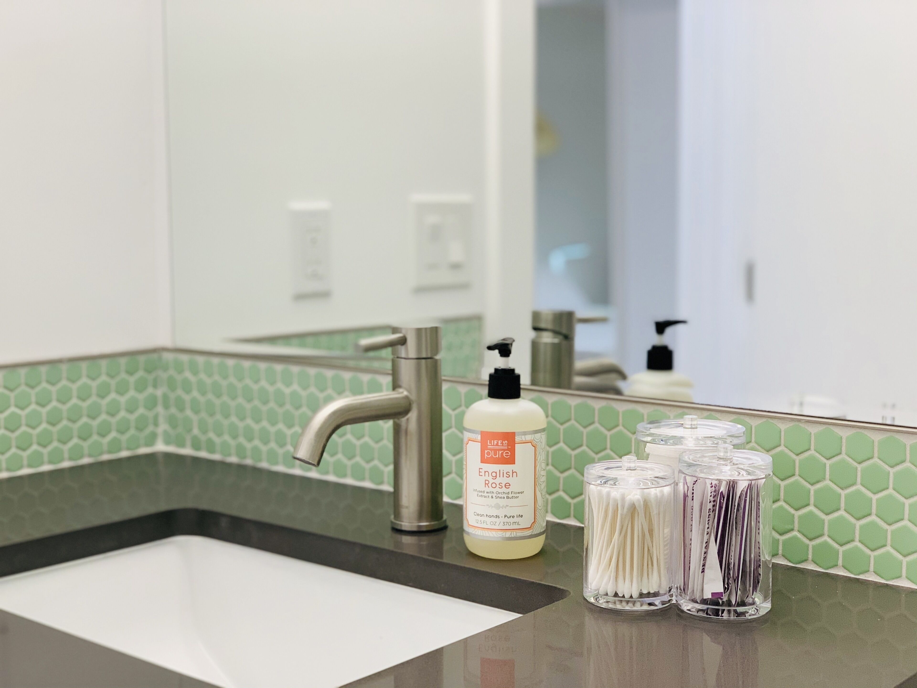 A bathroom sink with a green-tiled backsplash, a stainless steel faucet, and a bottle of hand soap.