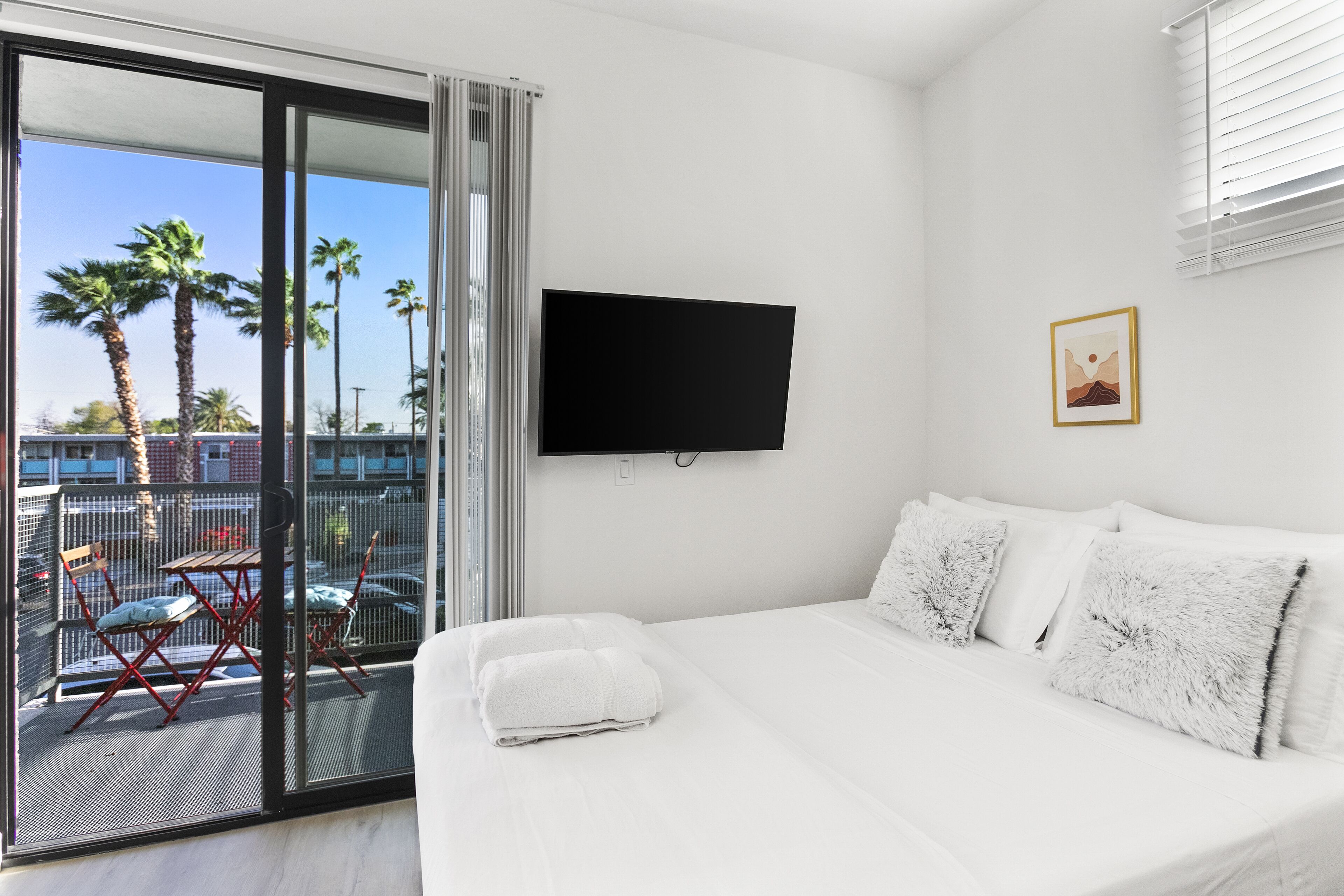 A hotel room with a bed, a TV mounted on the wall, a balcony with chairs, and a view of palm trees.