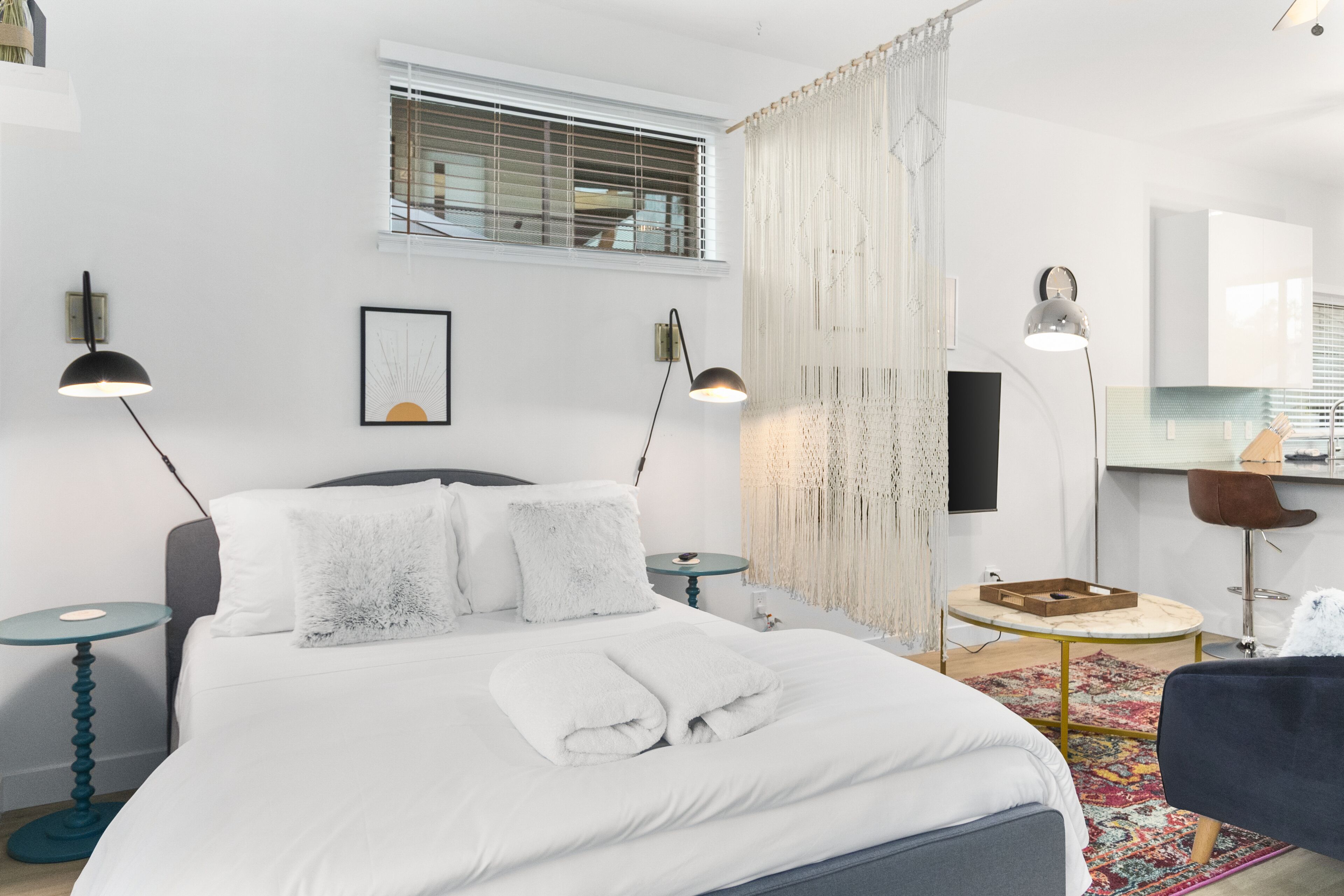 Una habitación de hotel moderna con una cama bien hecha, dos lámparas de noche, una mesita pequeña redonda, una silla y un espejo grande.