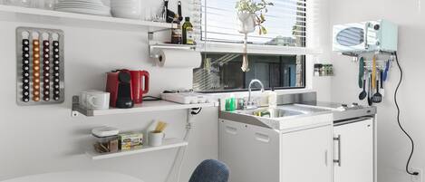 Une cuisine compacte avec des meubles blancs, un évier et un plan de travail. Des étagères au-dessus de l’évier servent à ranger la vaisselle et les bouteilles. On aperçoit une plante suspendue à travers la fenêtre.
