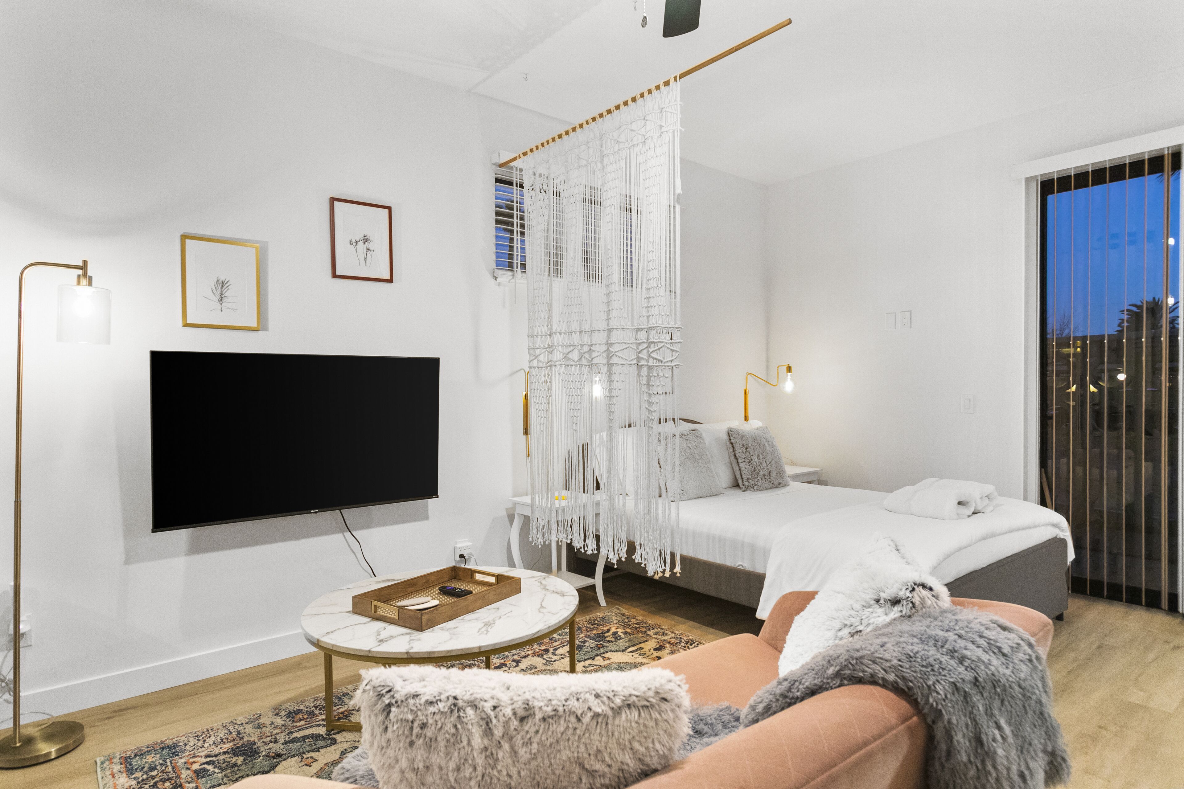 A modern bedroom with a bed, a TV, a decorative macrame wall hanging, and a large window.