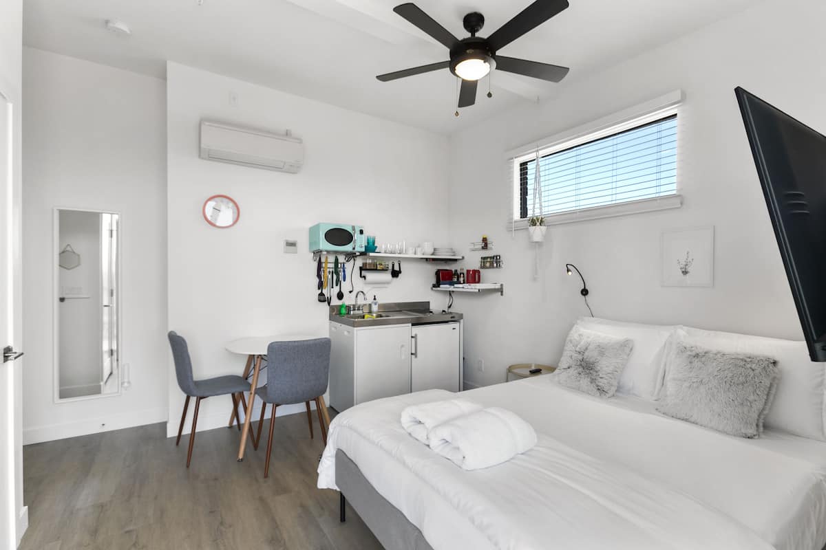 A compact hotel room with a bed, a desk, a chair, a ceiling fan, a window with blinds, and a small kitchenette.