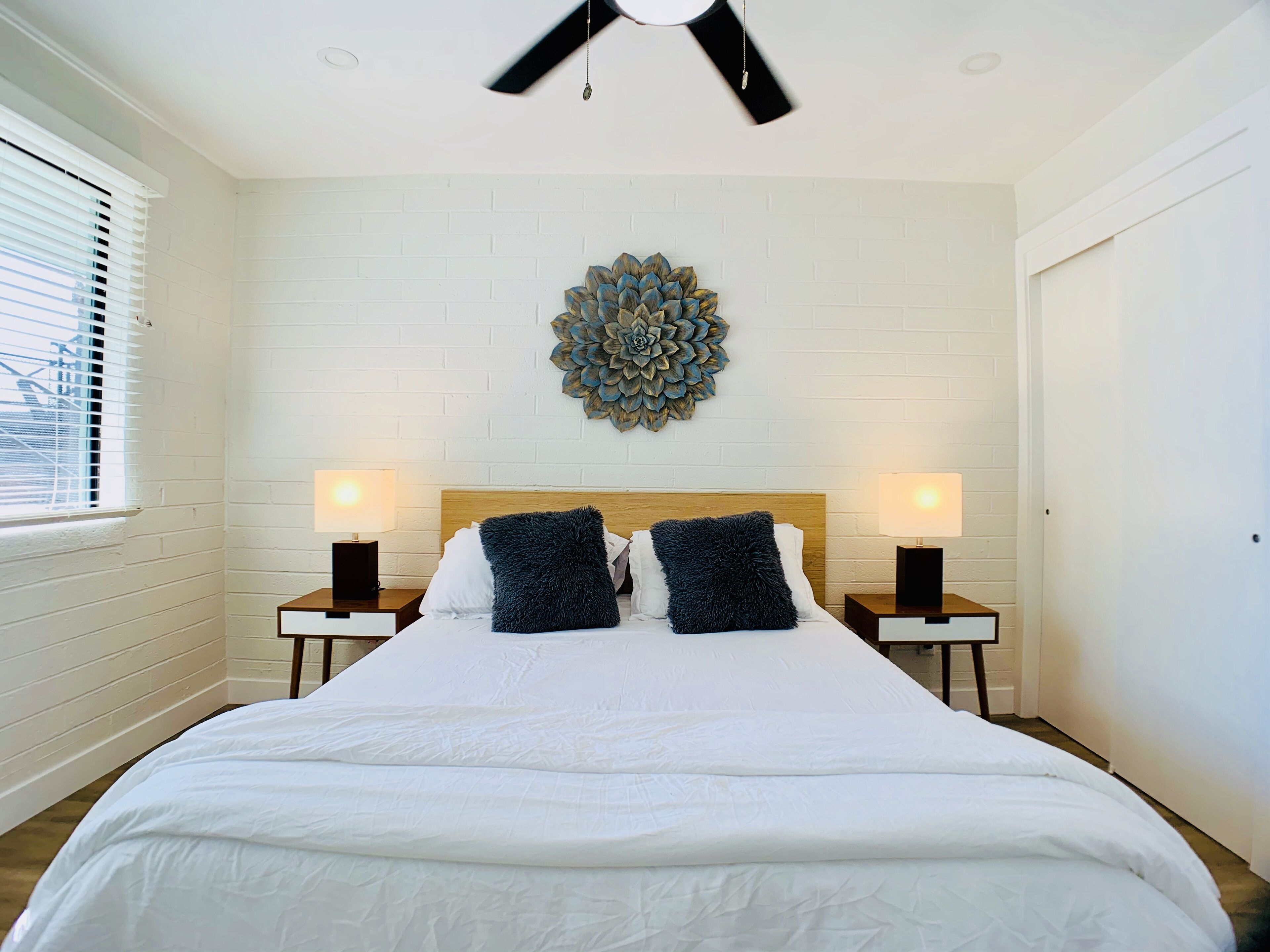 Quarto com cama grande, duas mesas de cabeceira com abajures, um detalhe decorativo na parede e um ventilador de teto.