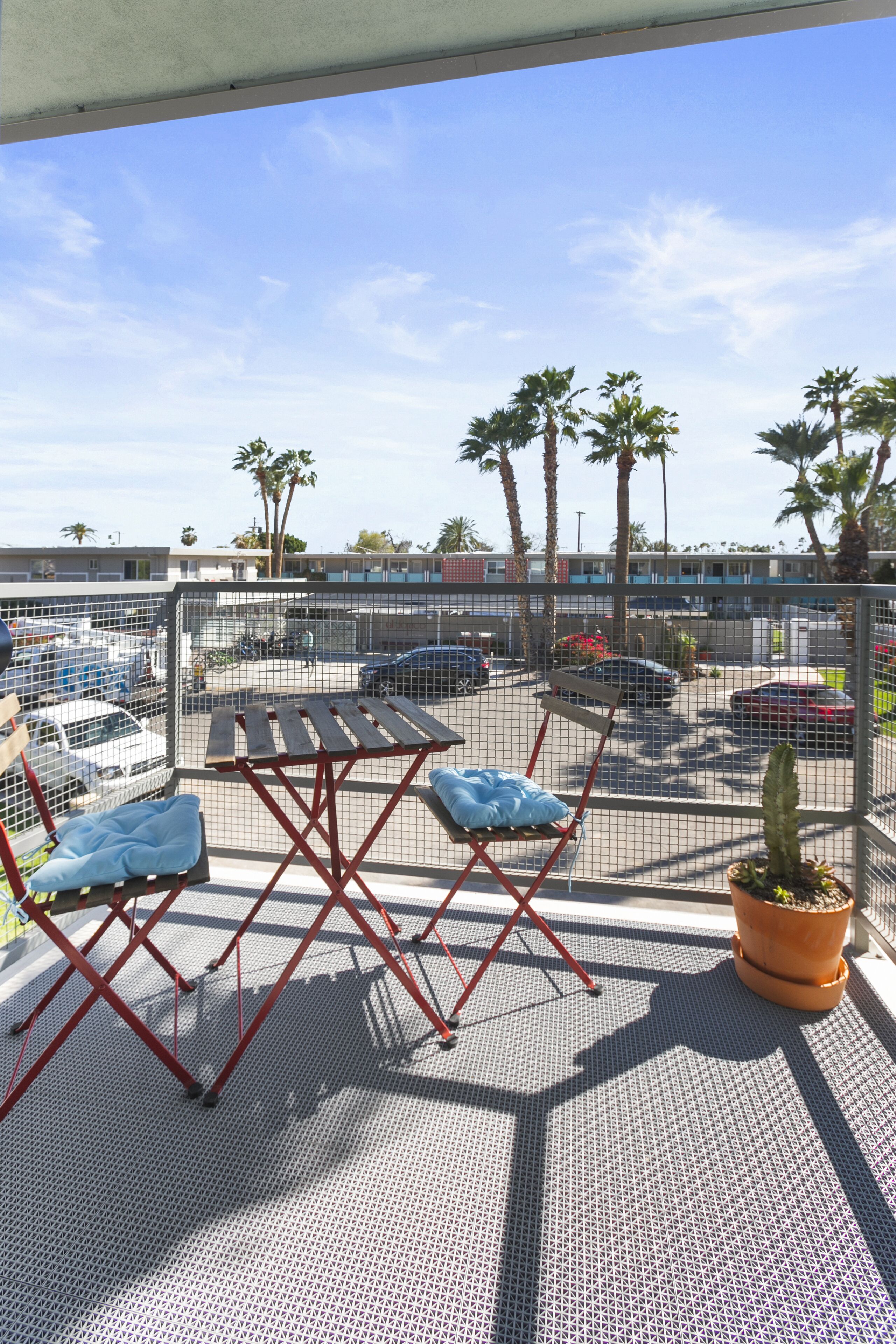 An outdoor seating area with a table and chairs, a potted plant, and a view of a parking lot and palm trees.